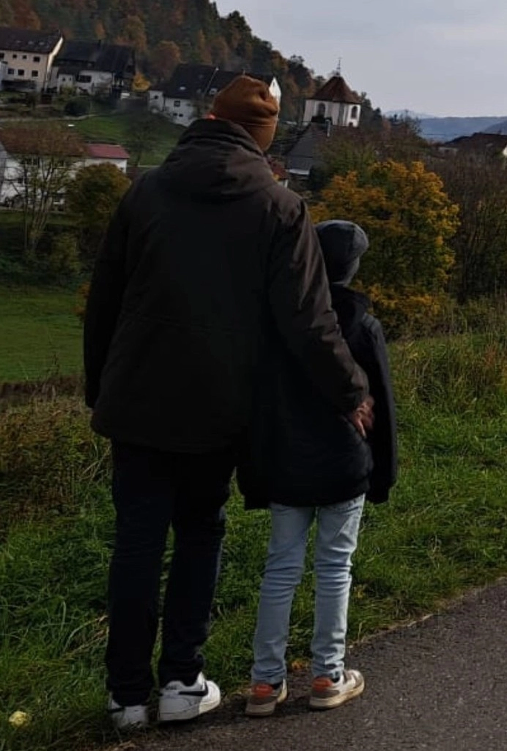 Spüren + Fühlen – Gesundheitskonzepte – zwei Personen, wahrscheinlich ein Elternteil und ein Kind, stehen auf einer Straße und blicken auf ein Dorf in einer hügeligen, ländlichen Gegend.